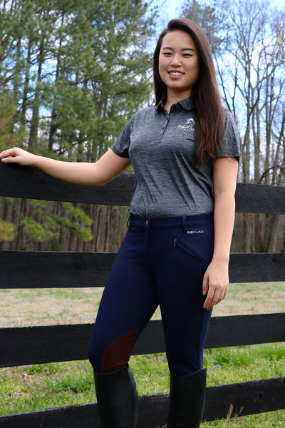 Gray Fitted Women's Polo Shirt with Buckwild Logo. Model in photo pairs navy blue and brown knee patch breeches from Spring 2017 lineup with gray polo while standing by a fence. 
