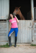 Model wears Women's Vintage Pink V-Neck Tee with Buckwild Logo with text "I like my horse better than most people" with royal blue and zebra print breeches while standingo utside a horse's stall.