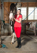 Model wears Red V-Neck Women's T-shirt with "Bitch Please, I Ride a Mare!" text on front paird with red riding breeches while standing next to her horse in a barn.